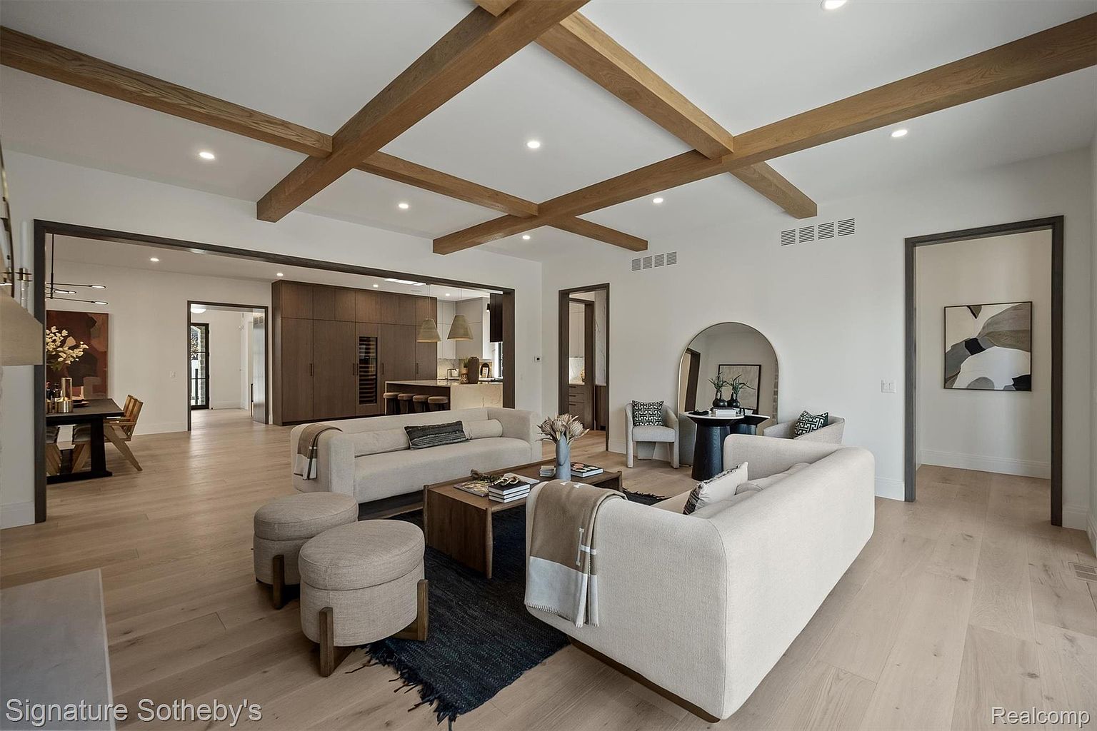 This is a spacious living room featuring hardwood floors, exposed wooden beams on the ceiling, and two light-colored sofas. A wooden coffee table sits on a dark rug, and there are two upholstered ottomans nearby. The room has a modern, open feel with doorways leading to other areas of the house, including the kitchen and dining area.