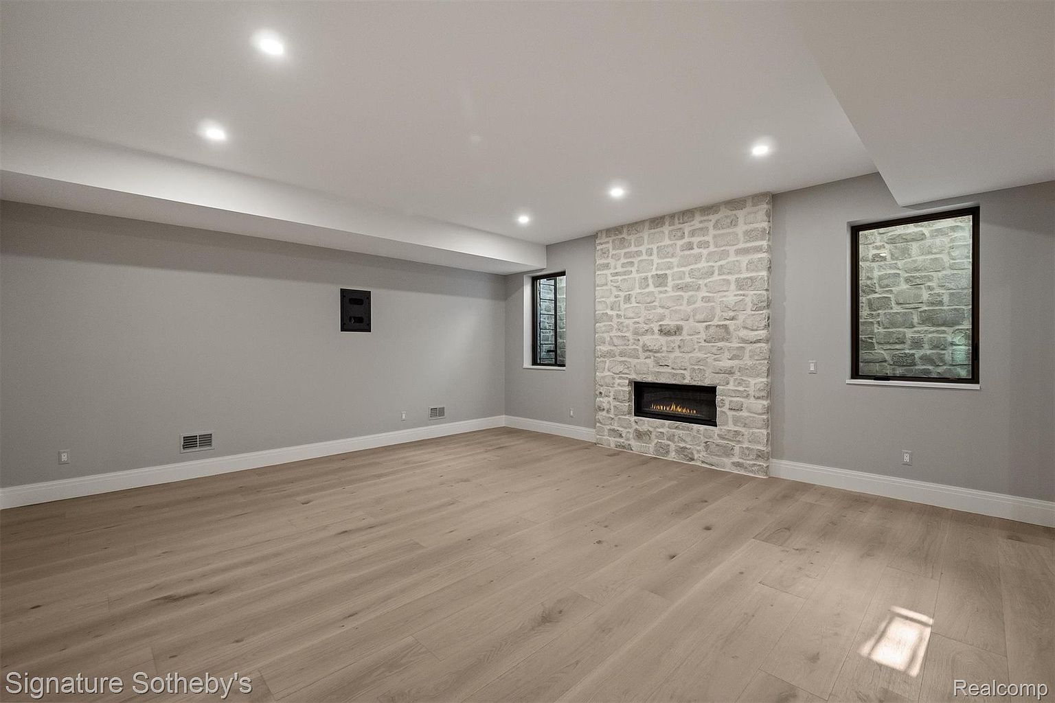 The image showcases a finished basement with light wood flooring and gray walls. A stone fireplace with a modern firebox serves as a focal point, flanked by windows that provide natural light. Recessed lighting illuminates the space, creating a clean and inviting atmosphere.