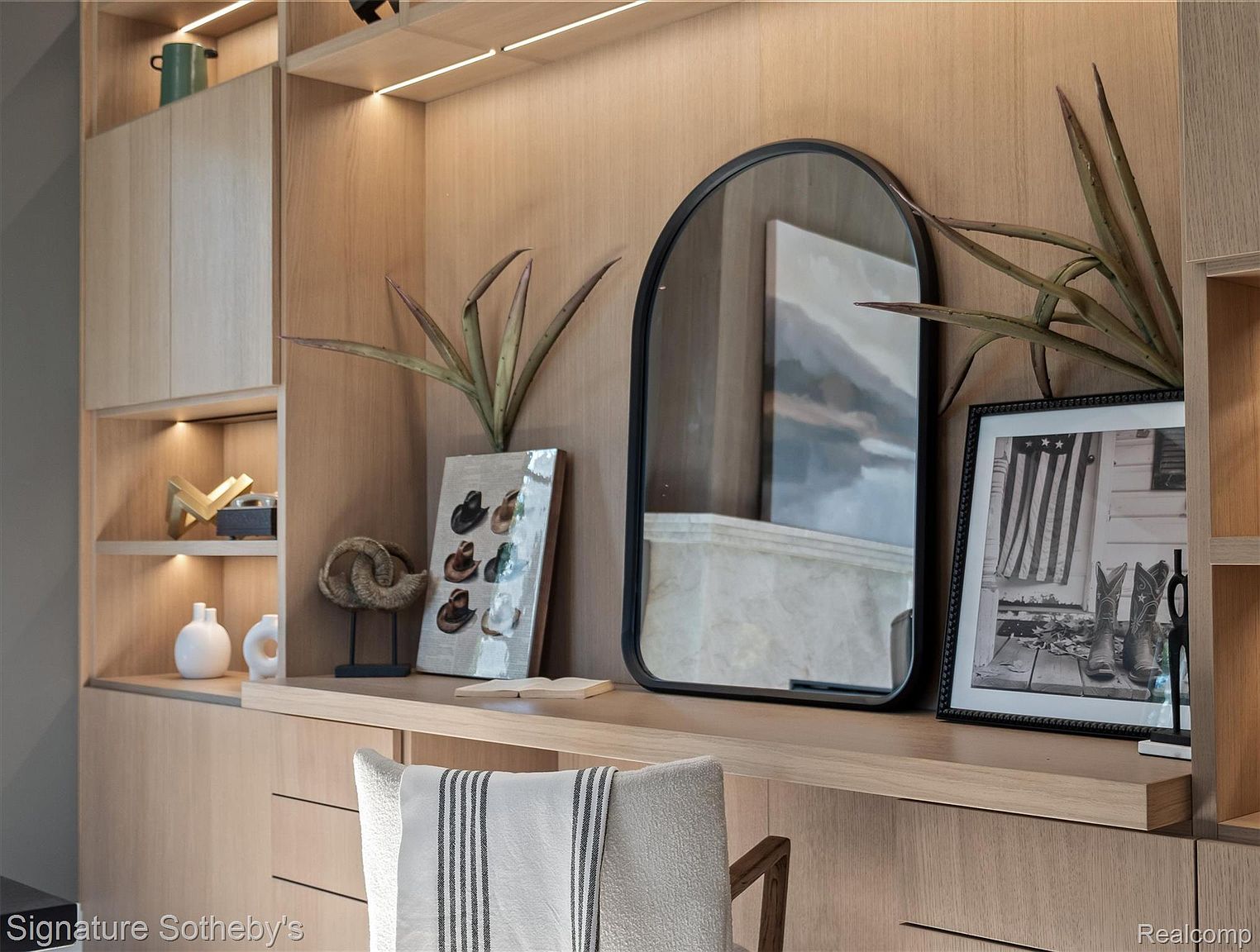 This image showcases a well-organized and stylish office or study area. The built-in shelving and cabinets, finished in a light wood tone, provide ample storage and display space. Decorative items such as a mirror, artwork, and plants add a touch of personality and sophistication to the room.