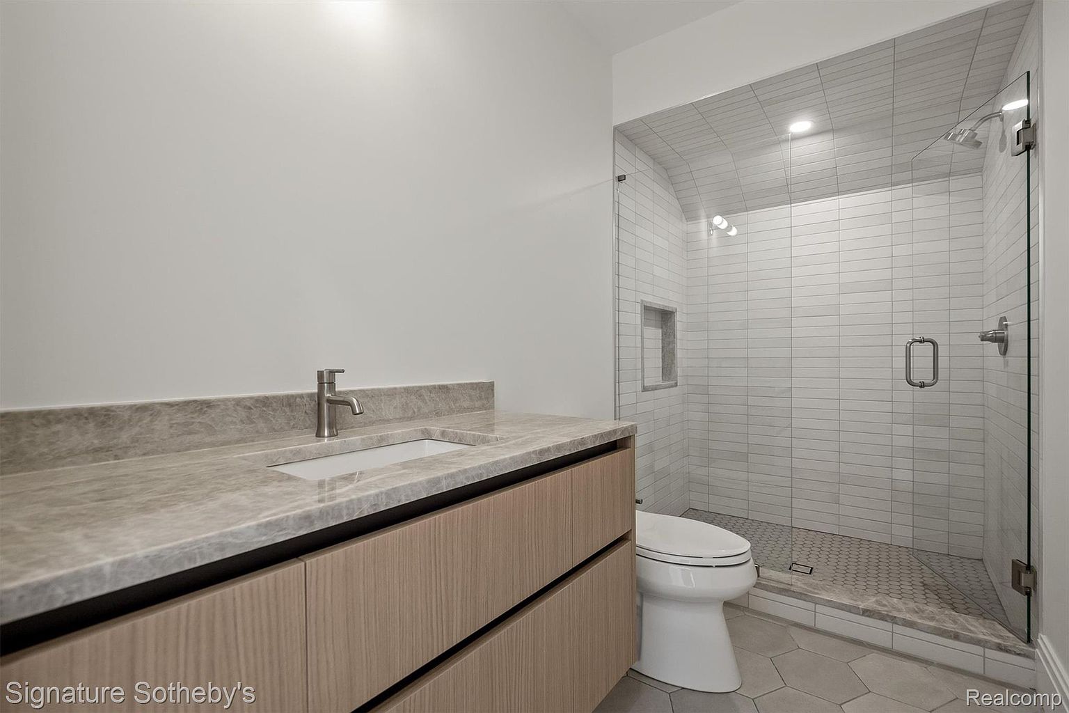 This is a modern bathroom featuring a floating vanity with a light wood finish and a gray marble countertop. A sleek, rectangular sink and a minimalist faucet complement the vanity. The shower is enclosed with glass and features white subway tile and a gray, hexagonal tile floor. A white toilet sits next to the shower, and the walls are painted a neutral gray.