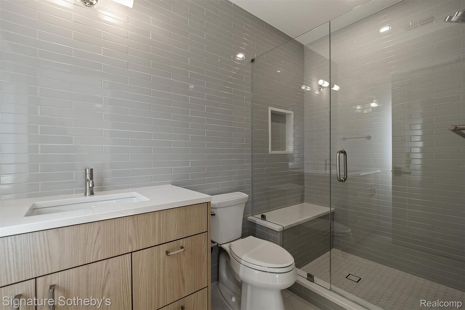 This is a modern bathroom featuring gray subway tile walls and a glass-enclosed shower. The vanity has a light wood finish with a white countertop and a rectangular sink. A toilet is positioned next to the shower, and the shower includes a built-in bench and a niche for toiletries. The overall impression is clean, contemporary, and well-lit.