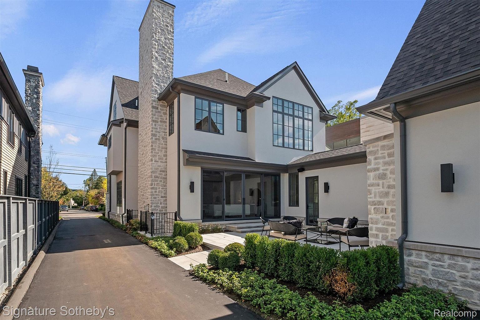 This is a rear exterior view of a modern luxury home. The house features a light-colored stucco exterior with dark trim around the windows and roofline. A stone chimney rises prominently, and a patio area with outdoor seating is visible, surrounded by manicured landscaping. The overall impression is one of sophisticated elegance and outdoor living.