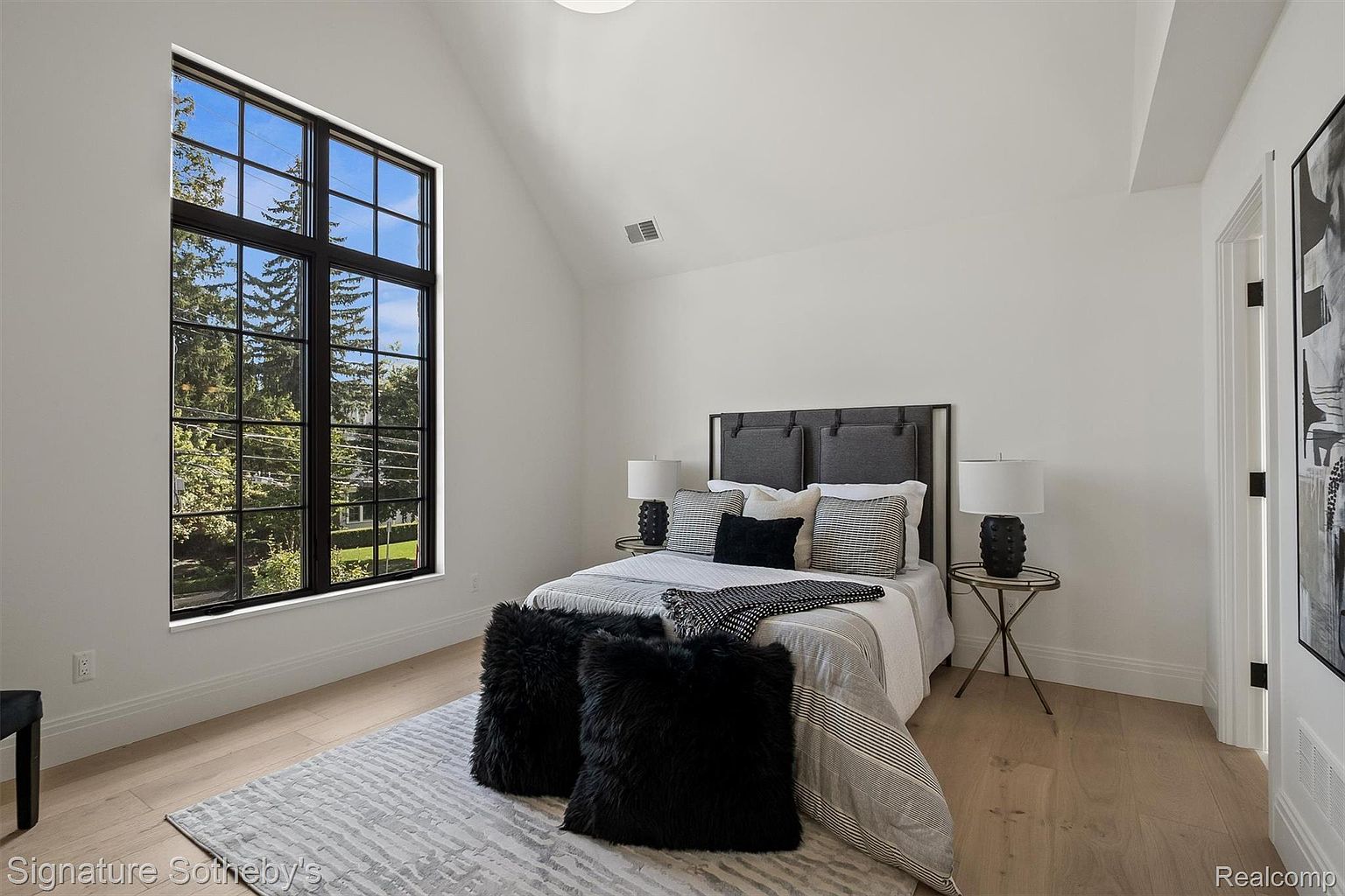 This is a bright and stylish bedroom featuring a large window that provides ample natural light and a view of the outdoors. The room is decorated in a modern style with a neutral color palette, accented by black and gray tones in the bedding and decor. The hardwood floors and minimalist furniture contribute to a clean and inviting atmosphere.
