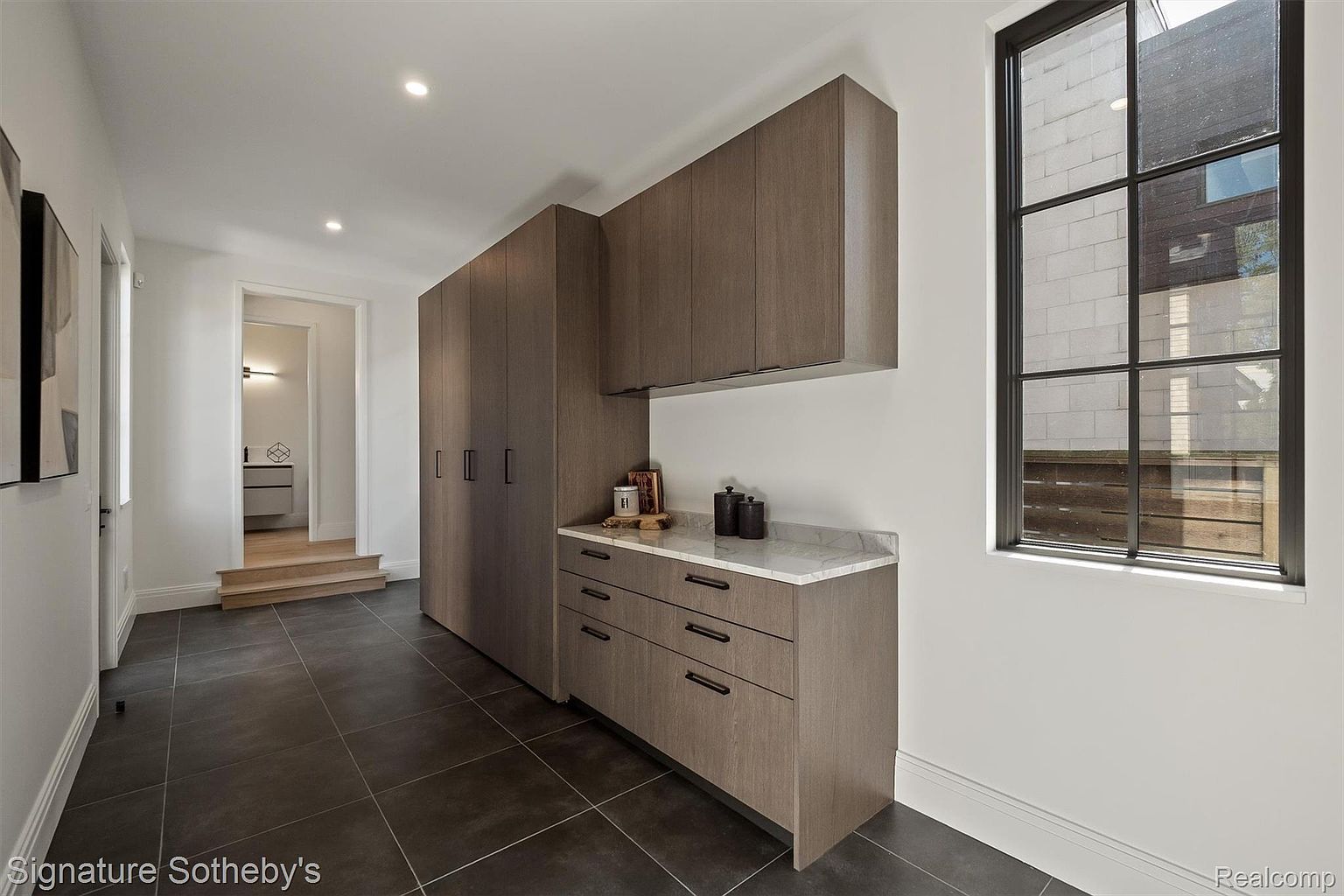 This interior shot showcases a modern hallway with dark tiled flooring and a built-in storage unit featuring sleek, handleless cabinets and drawers. A marble countertop provides a clean surface, while a large window with black framing allows natural light to flood the space. The hallway leads to a slightly elevated area, adding depth to the composition.