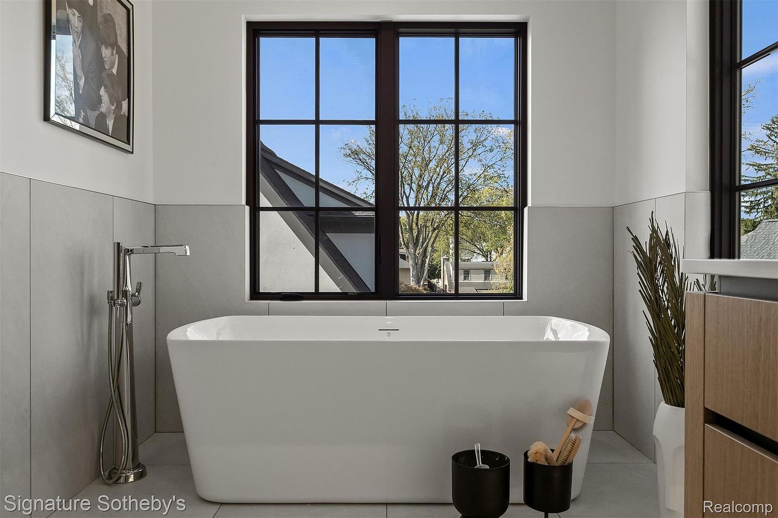 This is a bright and modern primary bathroom featuring a freestanding white bathtub positioned in front of a large window with dark frames, offering a view of the outdoors. The walls are a combination of white and light gray tiles, complemented by a sleek chrome faucet. The overall impression is clean, luxurious, and spa-like.