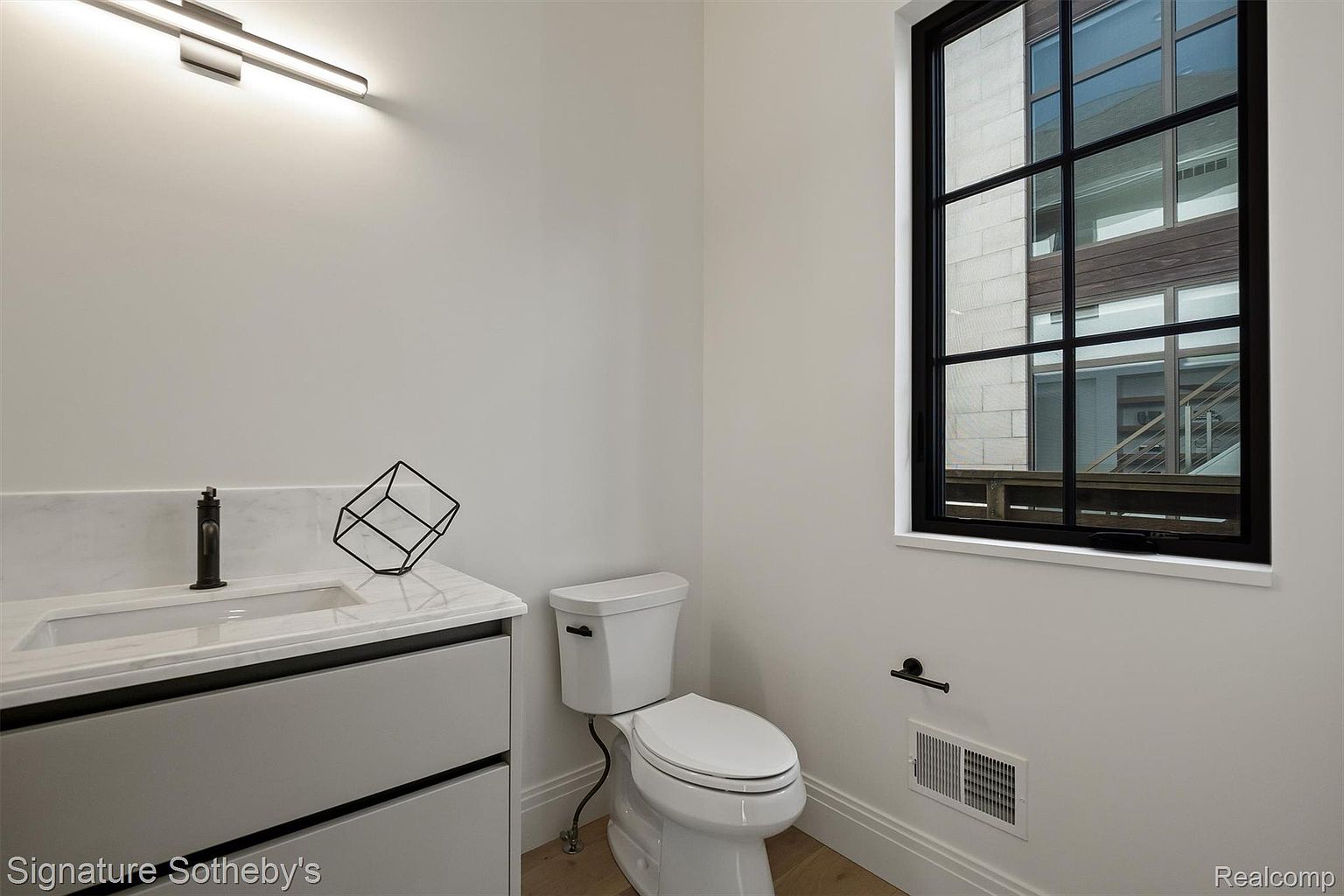 This is a well-lit guest bathroom featuring a modern vanity with a marble countertop and a sleek, dark faucet. A contemporary black wire sculpture sits on the counter, adding a touch of artistic flair. The room also includes a toilet and a window with a black frame, offering a view of the outside.