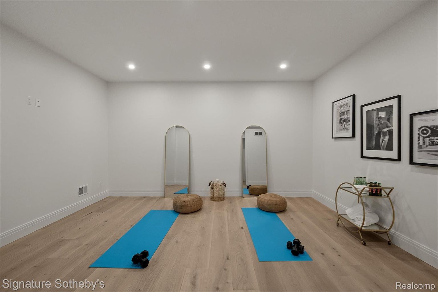 This is an interior shot of a home gym featuring light wood flooring, white walls, and recessed lighting. The room is equipped with two blue yoga mats, pairs of dumbbells, and round woven poufs. Two arched mirrors are placed against the wall, and framed black and white photographs adorn the right side of the room, along with a gold bar cart.