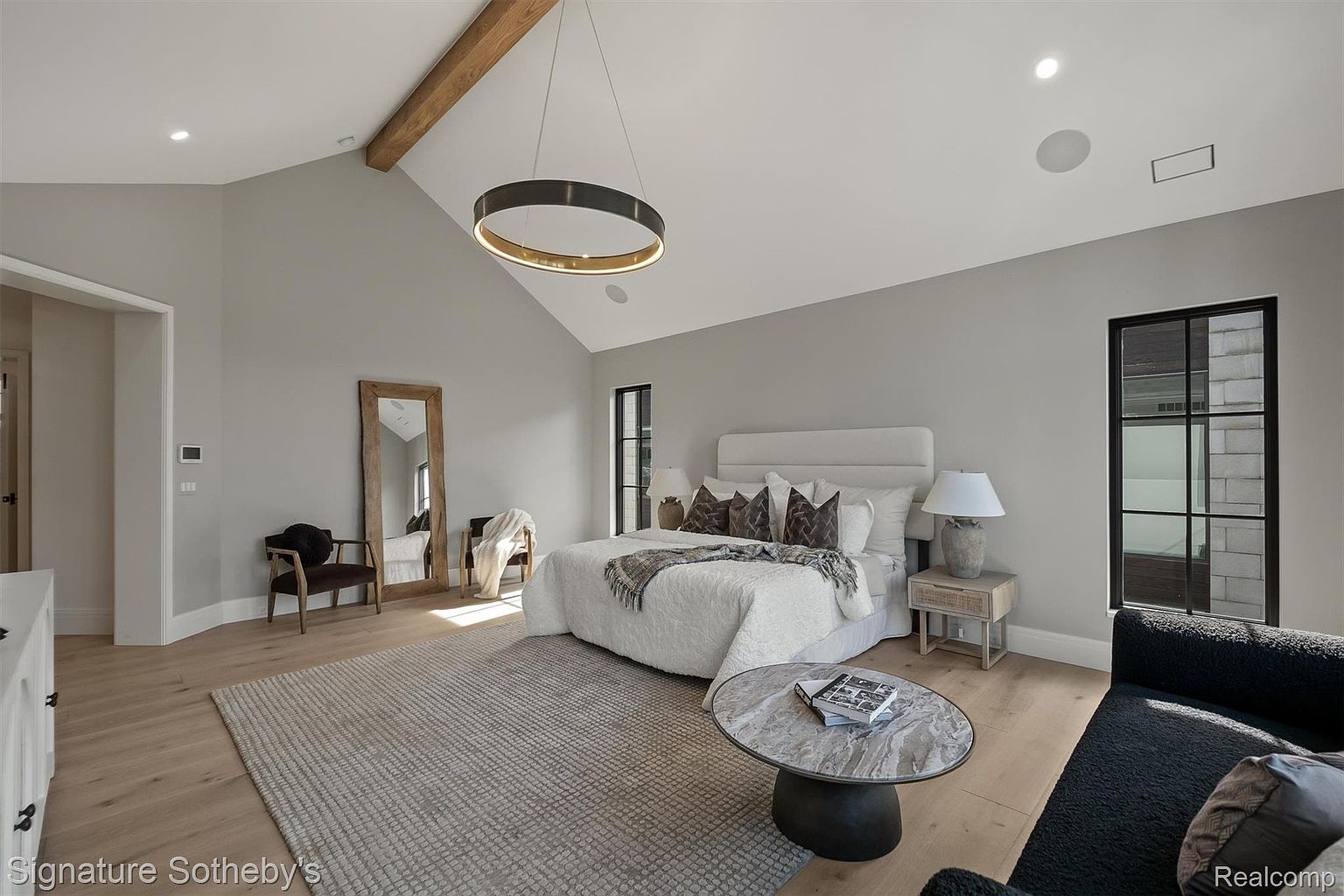 This is a primary bedroom featuring a neutral color palette with gray walls and light wood flooring. The room includes a large bed with a white headboard and textured bedding, a modern circular chandelier, and a full-length mirror. The space is well-lit with natural light from a window and recessed lighting, creating a serene and inviting atmosphere.
