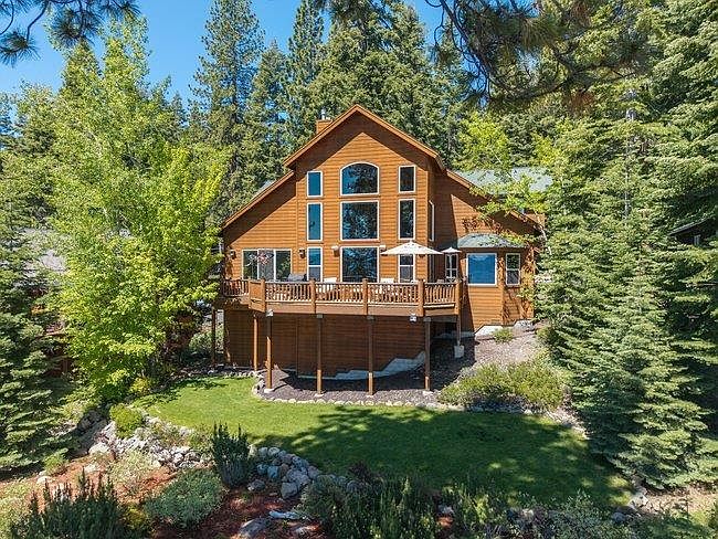 This rear view of a multi-story wooden home showcases a large, elevated deck that spans the width of the house, providing an ideal space for outdoor entertaining. The structure features a prominent, symmetrical arrangement of large windows that allow for abundant natural light and views of the surrounding forest. Nestled within a lush, tree-filled landscape, the property includes a well-maintained lawn and a stone-bordered garden area, creating a serene and private mountain retreat atmosphere.