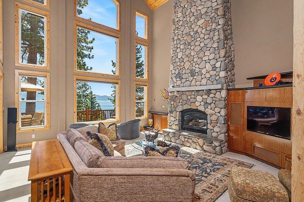 This spacious living room features a prominent stone-clad chimney breast that serves as a focal point, complemented by a large television integrated into the adjacent wood cabinetry. Tall windows provide an expansive view of the lake and surrounding trees, bathing the room in natural light. The space is furnished with a comfortable sofa and patterned area rug, creating a warm and inviting atmosphere.