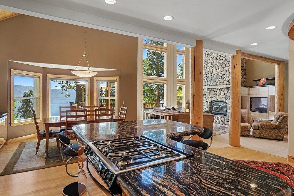 This open-concept kitchen features a prominent island with a dark, multi-toned stone countertop and an integrated gas cooktop. The space flows seamlessly into a dining area with large windows overlooking a lake and a living room anchored by a stone-clad chimney breast. Natural light floods the interior, highlighting the warm wood tones and the rustic, lodge-inspired architectural elements like the exposed timber columns.