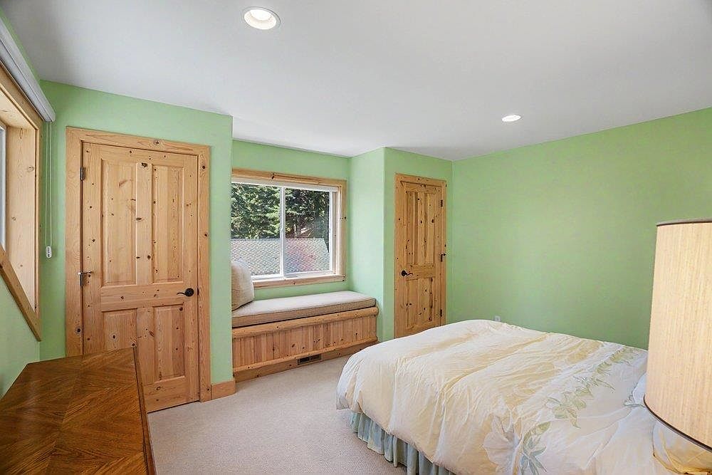 This bedroom features light green walls, neutral carpeting, and two natural wood doors that provide a rustic aesthetic. A built-in window seat with a cushioned top sits beneath a window, offering a cozy nook with a view of the outdoors. The room is furnished with a bed dressed in light-colored bedding and is illuminated by recessed ceiling lights.