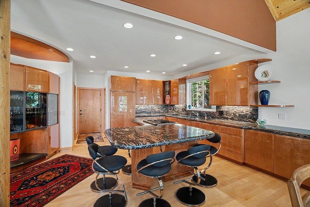 This kitchen features high-gloss wood cabinetry and a prominent island with a dark, patterned stone countertop, providing ample seating with four modern bar stools. The space is illuminated by recessed lighting and includes a window above the sink that looks out onto a wooded area. A rich, patterned rug adds a touch of color to the light wood flooring, creating a warm and inviting atmosphere.