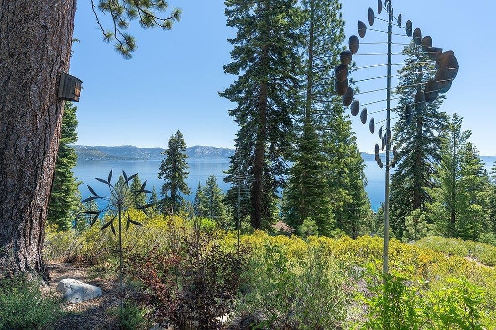 This scenic outdoor space features a lush, sloping garden filled with vibrant yellow wildflowers and mature pine trees. Two unique metal kinetic sculptures stand prominently in the foreground, while a birdhouse is mounted on a large tree trunk to the left. The background offers a breathtaking, expansive view of a deep blue lake framed by distant mountain ranges under a clear sky.