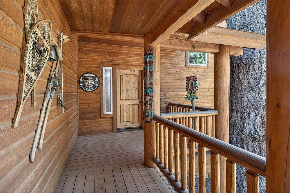 This rustic covered porch features warm wood-paneled walls and a matching wooden door, creating a cozy, cabin-like atmosphere. The space is decorated with traditional snowshoes and a circular indigenous-style art piece, while a carved wooden pillar adds a unique architectural detail. A wooden railing overlooks the surrounding forest, with a large tree trunk integrated into the deck structure, emphasizing a seamless connection to the natural environment.