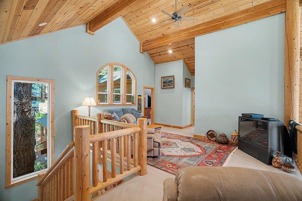 This rustic-style living area features a vaulted ceiling with exposed light wood beams and paneling, creating a warm, cabin-like atmosphere. The space includes a lofted landing with a log-style railing overlooking the stairs, a patterned area rug, and a television console. Large windows, including an arched window, provide natural light and views of the surrounding trees, while the light blue walls offer a soft contrast to the natural wood elements.