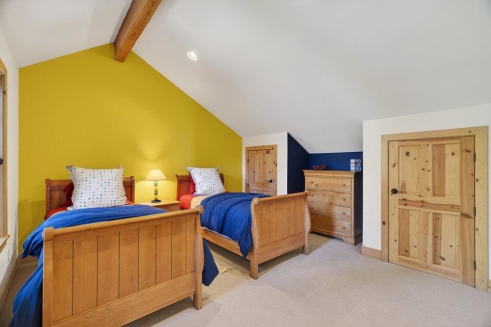 This bedroom features two twin-sized wooden beds with blue bedding and patterned pillows, set against a vibrant yellow accent wall. The room includes a wooden dresser, a bedside lamp, and light-toned carpeting, all under a vaulted ceiling with a exposed wooden beam. The space is bright and functional, with a wooden door leading to another area of the home.