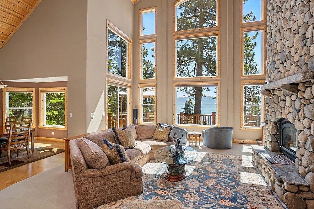 This spacious living room features a vaulted wood-paneled ceiling and a prominent stone-clad chimney breast that anchors the space. A large sectional sofa and a glass-topped coffee table are arranged on a patterned area rug, creating a comfortable seating area. Tall windows provide an expansive view of the outdoors, filling the room with natural light and highlighting the open-concept layout that flows into the adjacent dining area.