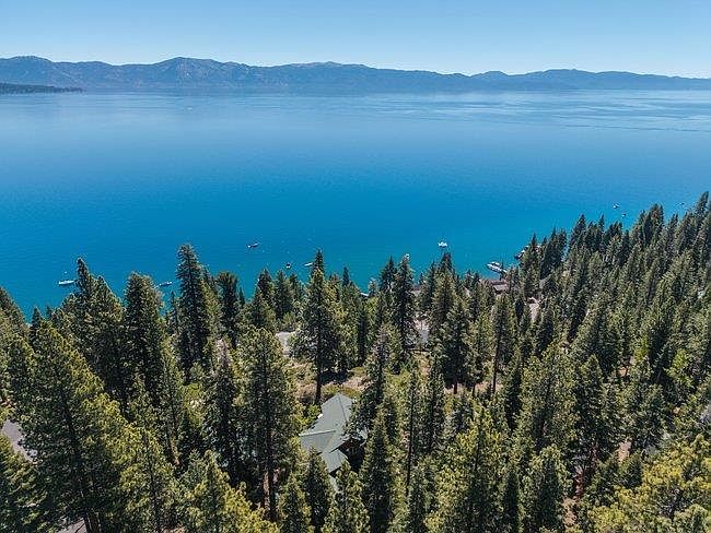 This high-angle aerial shot captures a secluded home nestled within a dense, lush forest of evergreen trees overlooking a vast, deep blue lake. The perspective emphasizes the property's serene, private setting and its direct proximity to the water, with several small boats visible on the lake surface. The clear, bright sky and expansive horizon create a tranquil and picturesque atmosphere, highlighting the home's integration into the natural landscape.