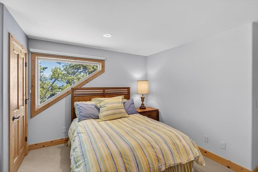 This cozy bedroom features a wooden bed frame centered against a light-colored wall, dressed in a striped comforter with matching pillows. A unique, angular window provides a view of the surrounding trees, while a bedside table with a lamp adds a warm touch to the space. The room is finished with light wood trim along the baseboards and a wooden door, creating a rustic and inviting atmosphere.