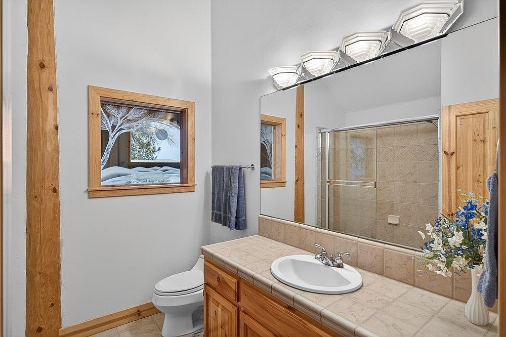 This bathroom features a rustic aesthetic with light wood trim, a tiled vanity countertop, and a matching tiled backsplash. A large mirror reflects the shower area, which is enclosed by glass doors and finished with neutral-toned wall tiles. The space is illuminated by a multi-light fixture above the mirror and includes a window that provides a view of a snowy outdoor scene.