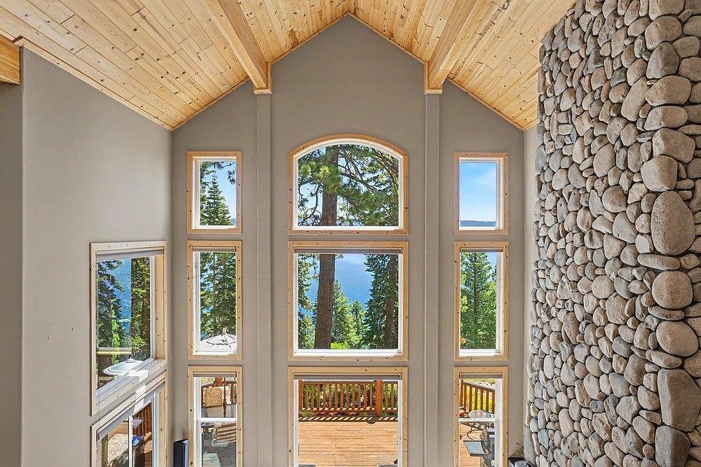 This interior view captures a grand living space with a vaulted ceiling finished in light wood planks. A massive stone-clad chimney breast dominates the right side of the room, while a series of tall, stacked windows provide expansive views of the surrounding forest and lake. The neutral gray walls and natural wood accents create a rustic yet airy atmosphere, emphasizing the height and architectural scale of the room.