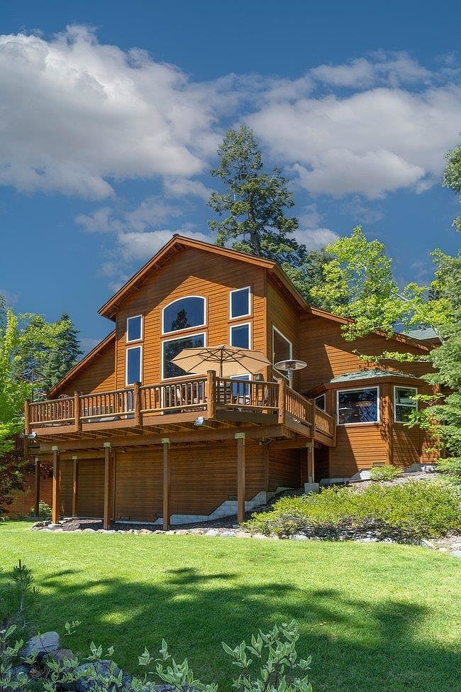 This image captures the rear exterior of a multi-level wooden home nestled among lush green trees under a bright, cloudy sky. A spacious elevated deck with a wooden railing extends from the main living area, featuring a large umbrella and outdoor seating, while a well-manicured lawn stretches out in the foreground. The home's design incorporates large windows and a warm, natural wood siding, creating a welcoming and serene mountain-retreat aesthetic.