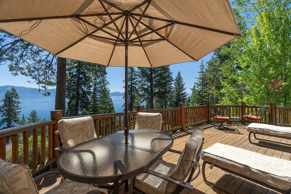 This elevated wooden deck offers a serene outdoor living space with a panoramic view of a large lake surrounded by dense pine forests. The area is furnished with a glass-topped dining table under a large tan umbrella and several lounge chairs, creating an inviting spot for relaxation. The perspective is from a slightly elevated vantage point, capturing the expansive natural scenery and the warm, sunny atmosphere of the deck.
