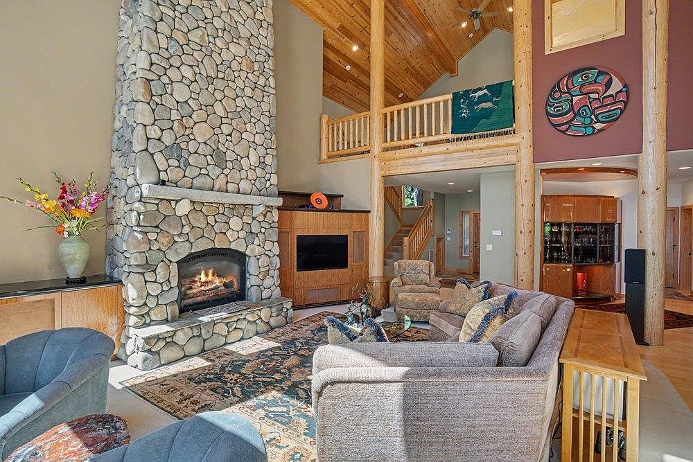 This spacious living room features a prominent stone-clad chimney breast that serves as the focal point of the space. The room boasts a vaulted wood-paneled ceiling, a lofted balcony area, and a large, patterned area rug anchoring the seating arrangement. The open-concept design flows into a secondary living area and a staircase, creating a warm, rustic atmosphere.