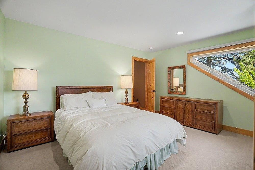 This bedroom features light green walls, a neutral carpet, and a queen-sized bed centered against the wall. A unique, angled window follows the roofline, allowing natural light to fill the space, while matching wooden nightstands and a dresser provide ample storage. The room is accented by warm wood trim around the door and window, creating a cozy and inviting atmosphere.