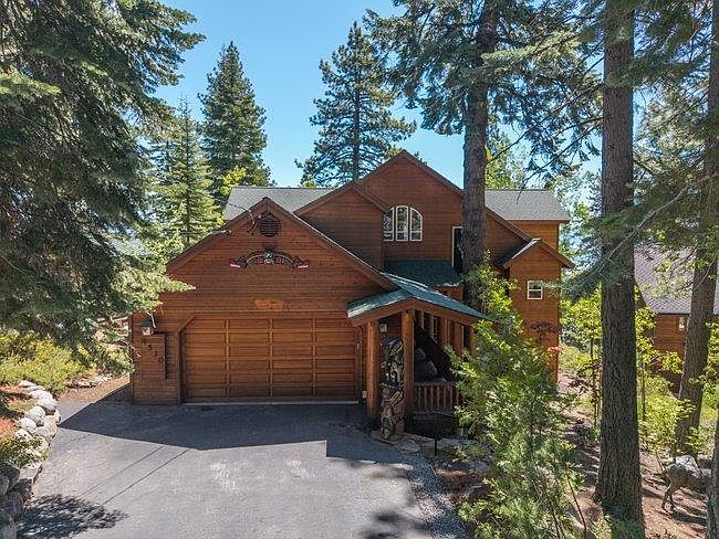 This two-story mountain-style home features warm wood siding and a prominent attached garage, nestled within a lush, forested landscape. A covered entryway is accented by a decorative carved wooden post, while the driveway provides ample parking space. The home's rustic aesthetic is complemented by the surrounding tall pine trees and natural, wooded setting.