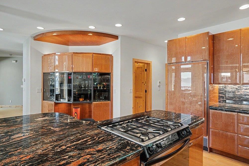 This kitchen features high-gloss, warm-toned wood cabinetry and a prominent island topped with dark, heavily veined stone. A gas cooktop is integrated into the island, while a built-in refrigerator matches the surrounding cabinetry. To the left, a custom-built, curved display cabinet with glass shelving adds a unique architectural element to the space.
