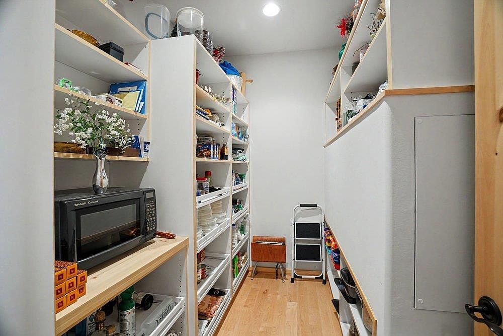 This organized walk-in pantry features extensive light wood shelving on both sides, providing ample storage space for kitchen essentials and dry goods. A black microwave sits on a dedicated shelf, while a small step ladder and a wooden crate are positioned at the end of the narrow aisle. The space is illuminated by recessed lighting, creating a bright and functional storage area.