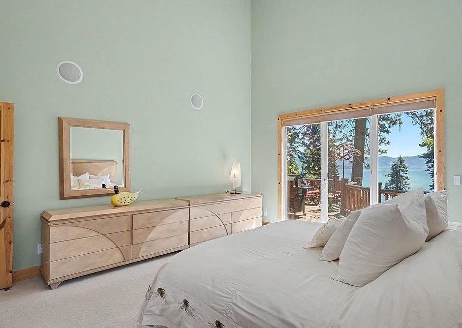 This bright and airy primary bedroom features light green walls and a large bed with white bedding, positioned to take advantage of the expansive view through the sliding glass doors. A long, light wood dresser with a matching mirror provides ample storage, while the room's high ceilings enhance the sense of space. The sliding doors open onto a deck, offering a scenic view of the surrounding trees and water.