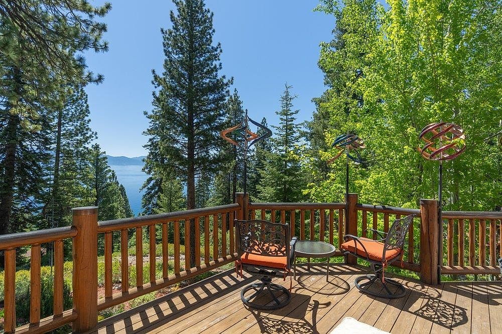 This elevated wooden deck offers a serene outdoor living space with a rustic log-style railing and a panoramic view of a lake surrounded by dense pine forests. The area is furnished with two comfortable swivel chairs and a small glass-topped table, complemented by decorative metal wind spinners. The perspective captures the inviting atmosphere of a mountain retreat, emphasizing the connection between the private deck and the natural landscape.