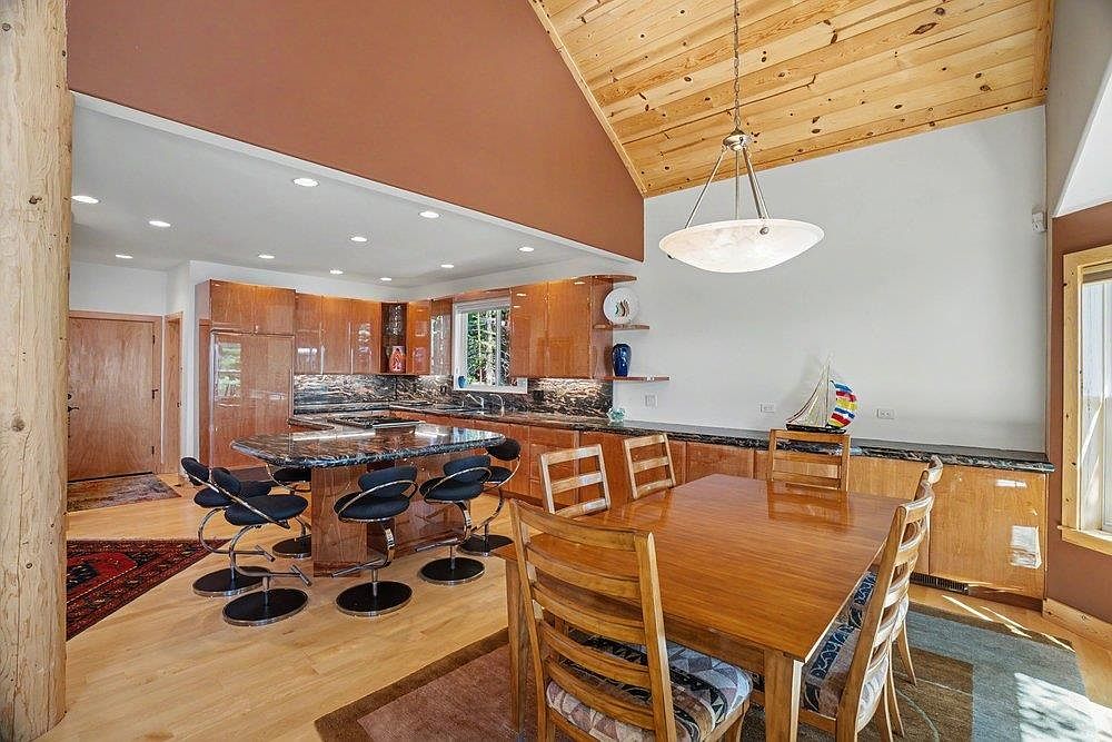 This open-concept kitchen and dining area features a vaulted wood-paneled ceiling and warm-toned cabinetry. A central island with a dark stone countertop provides bar seating, while a wooden dining table sits adjacent to the kitchen workspace. The space is illuminated by a large pendant light and recessed lighting, creating a cozy and inviting atmosphere.