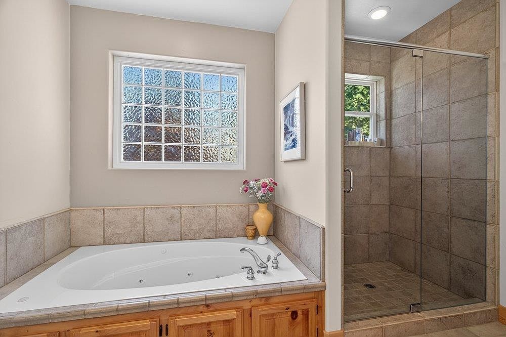 This bathroom features a jetted soaking tub set into a light wood-toned vanity base with a tiled surround. Adjacent to the tub is a walk-in shower enclosed by a glass door, featuring tiled walls and a small window. The space is accented by a glass block window above the tub and a framed piece of wall art, creating a functional and neutral bathing area.
