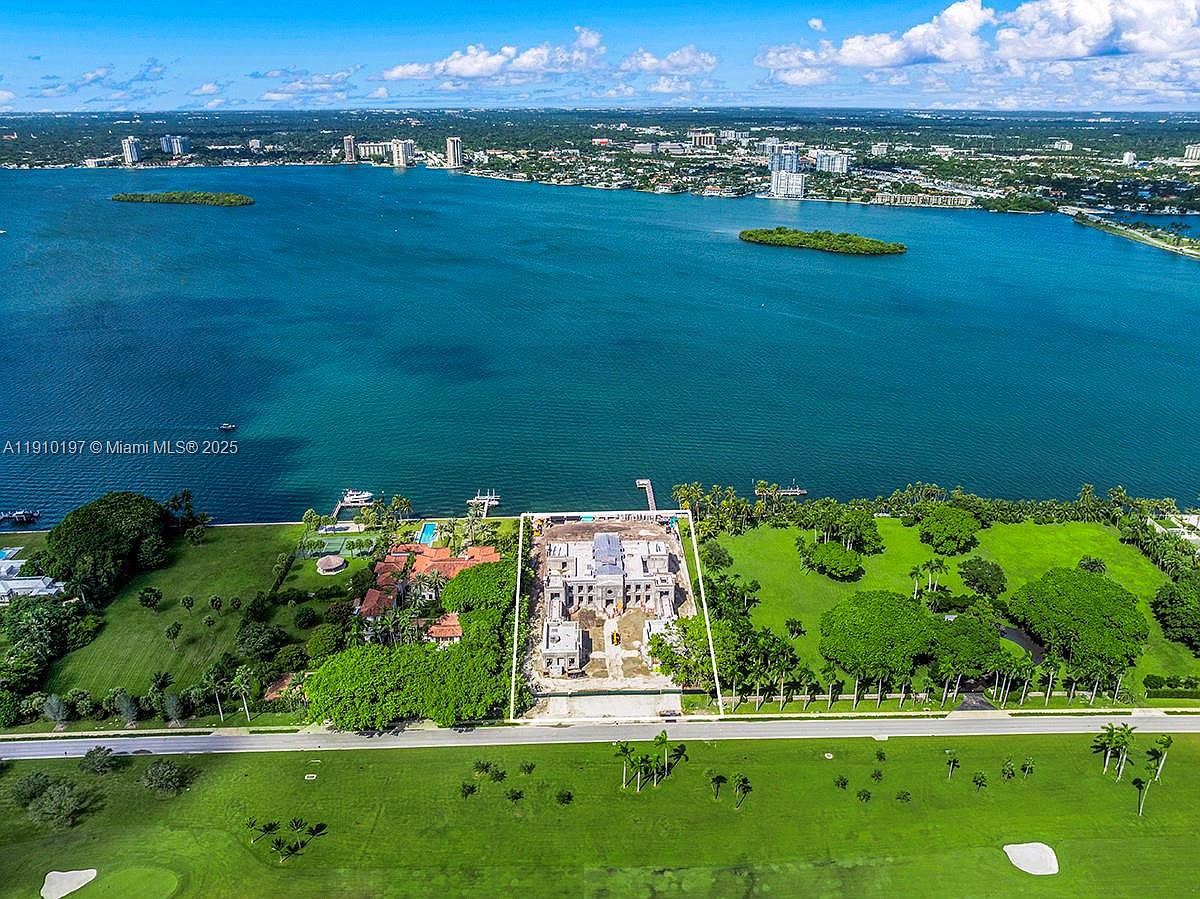This aerial shot showcases a grand estate under construction, bordered by lush greenery and waterfront views. The property features a large building footprint, suggesting a spacious layout, and is surrounded by manicured lawns and mature trees. The waterfront location adds significant appeal, promising stunning views and potential for water activities.