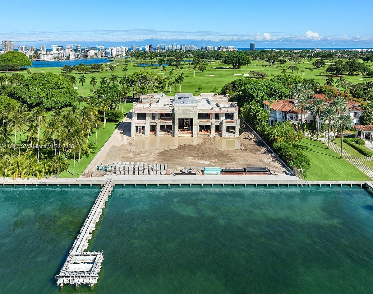 This aerial view showcases a waterfront property under construction, featuring a modern architectural design with a concrete structure. A long pier extends into the clear blue water, and the property is surrounded by lush greenery, including palm trees and a golf course. The cityscape is visible in the background, adding to the property's desirable location.