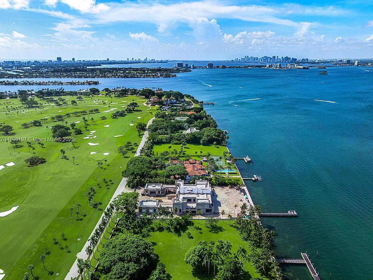 This aerial view showcases a luxurious waterfront property featuring a large modern mansion under construction, expansive green lawns, and a private dock extending into the turquoise water. The property is adjacent to a golf course and offers stunning views of the city skyline in the distance, creating an impression of exclusivity and high-end living.