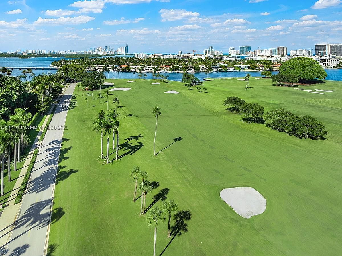 This aerial view showcases a meticulously maintained golf course with lush green fairways, strategically placed sand traps, and mature palm trees lining the course. In the background, a cityscape is visible across the water, adding a touch of urban sophistication to the serene landscape. A road runs along the edge of the golf course, providing access and enhancing the overall aesthetic appeal.