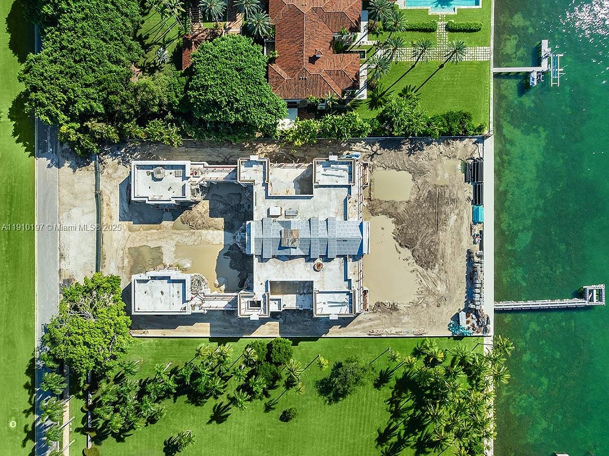 This aerial shot showcases a large property under construction, featuring a substantial building with a gray roof and multiple sections. The surrounding landscape includes lush green lawns, mature trees, and waterfront access with a dock. The image highlights the scale and prime location of the real estate.