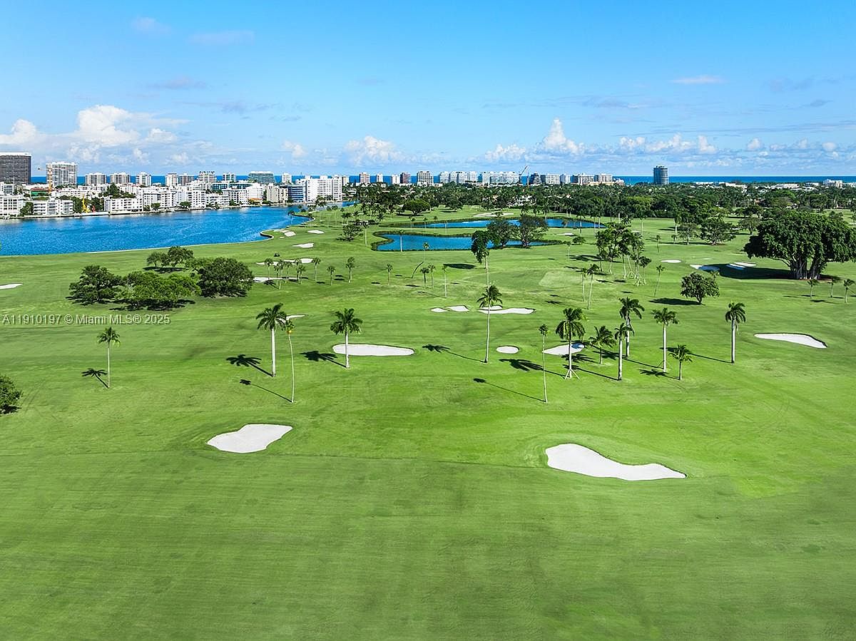 This aerial view showcases a lush green golf course with strategically placed sand traps and palm trees. A body of water borders the course, with a cityscape visible in the background under a clear blue sky with scattered clouds. The image conveys a sense of luxury and recreation, highlighting the property's proximity to both nature and urban amenities.