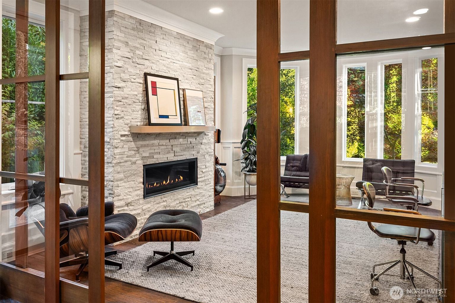 This sophisticated living space is viewed through a set of wooden-framed glass doors, creating a sense of depth and architectural interest. The room features a prominent stacked-stone chimney breast with a linear fireplace and a minimalist mantel, complemented by a plush area rug and mid-century modern lounge seating. Large windows line the far wall, bathing the room in natural light and providing a serene view of the surrounding greenery.