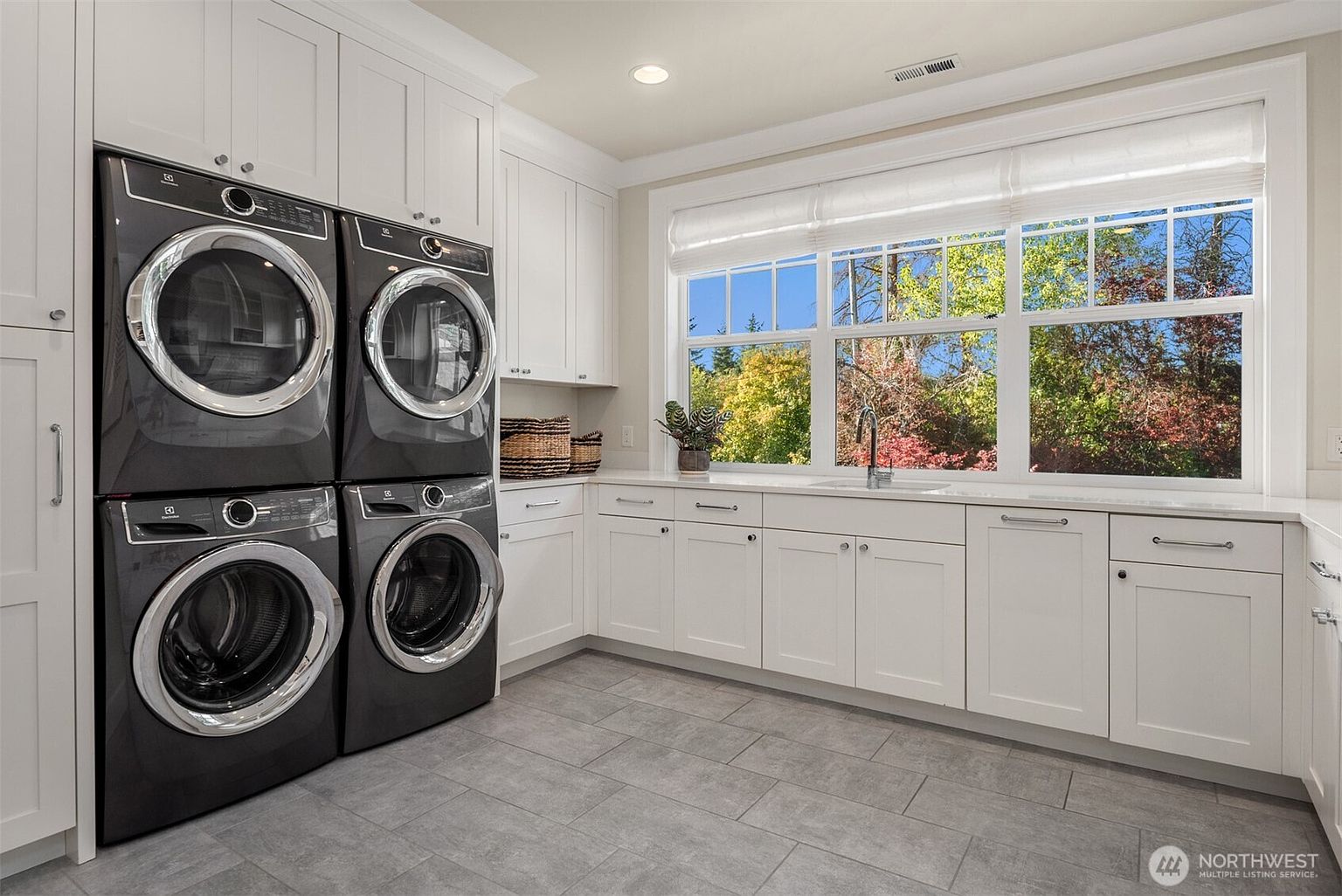 This bright and functional laundry room features a stacked washer and dryer set in a dark finish, contrasted against crisp white cabinetry and light-colored countertops. A large window above the sink provides ample natural light and a view of the outdoors, while the neutral tile flooring adds a clean, polished aesthetic. The space is well-organized with plenty of storage, creating a practical and inviting utility area.