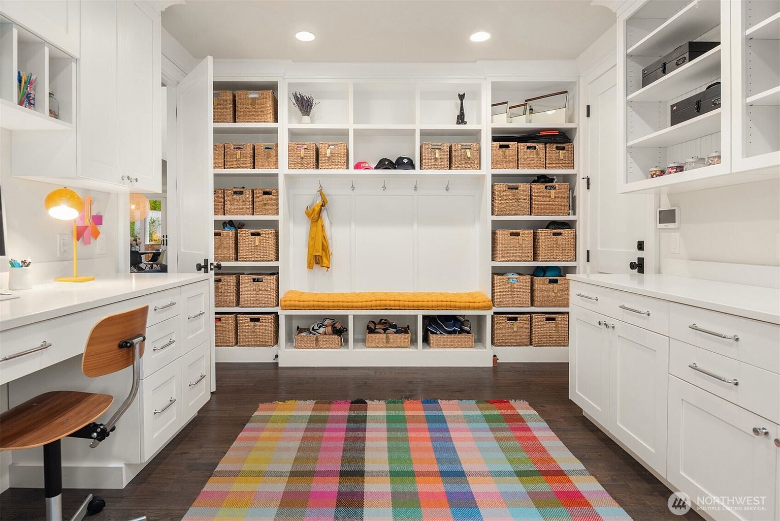 This organized mudroom and laundry area features extensive white built-in cabinetry with numerous woven storage baskets and a central bench with a yellow cushion. A colorful plaid rug anchors the space, which includes a desk area on the left and additional counter space on the right. The room is brightly lit by recessed ceiling lights, creating a clean and highly functional transition space.
