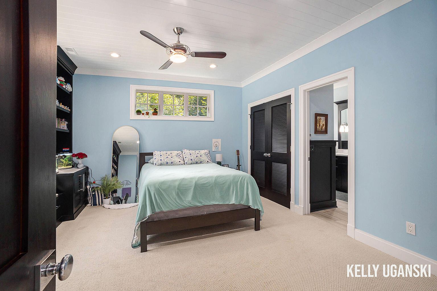This is a cozy bedroom featuring light blue walls, white trim, and a white plank ceiling with a ceiling fan. The room is furnished with a bed covered in a light green blanket, a dark wood dresser with a bookshelf, and a full-length mirror. There is a window above the bed and a closet with louvered doors to the right of the bed. The room has a calm and inviting atmosphere.