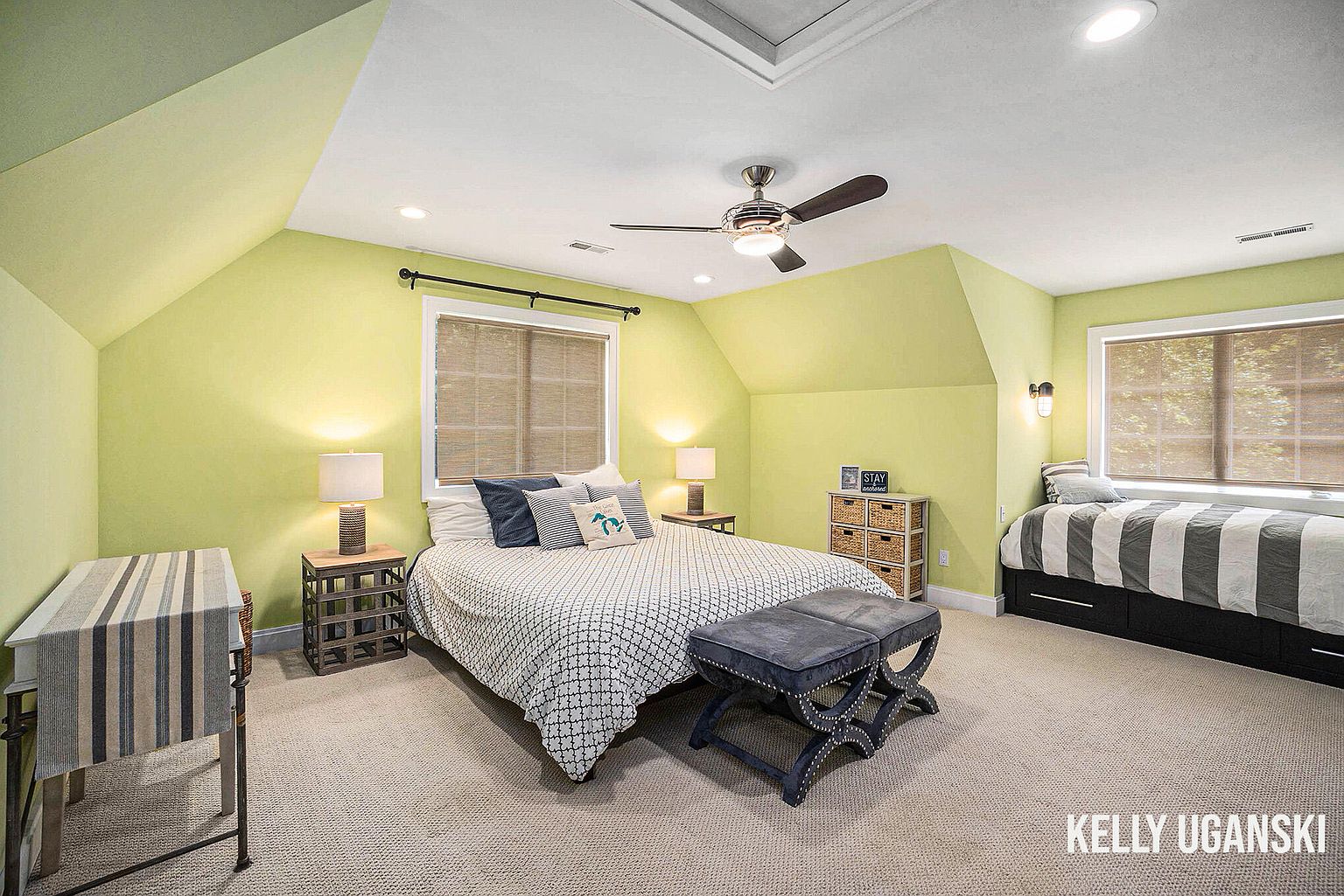 This is a guest bedroom featuring a bed with a patterned comforter, several decorative pillows, and a dark blue upholstered bench at the foot of the bed. The room has light green walls, carpeted floors, and two windows with woven shades. There is also a twin bed with a striped comforter on the right side of the room.