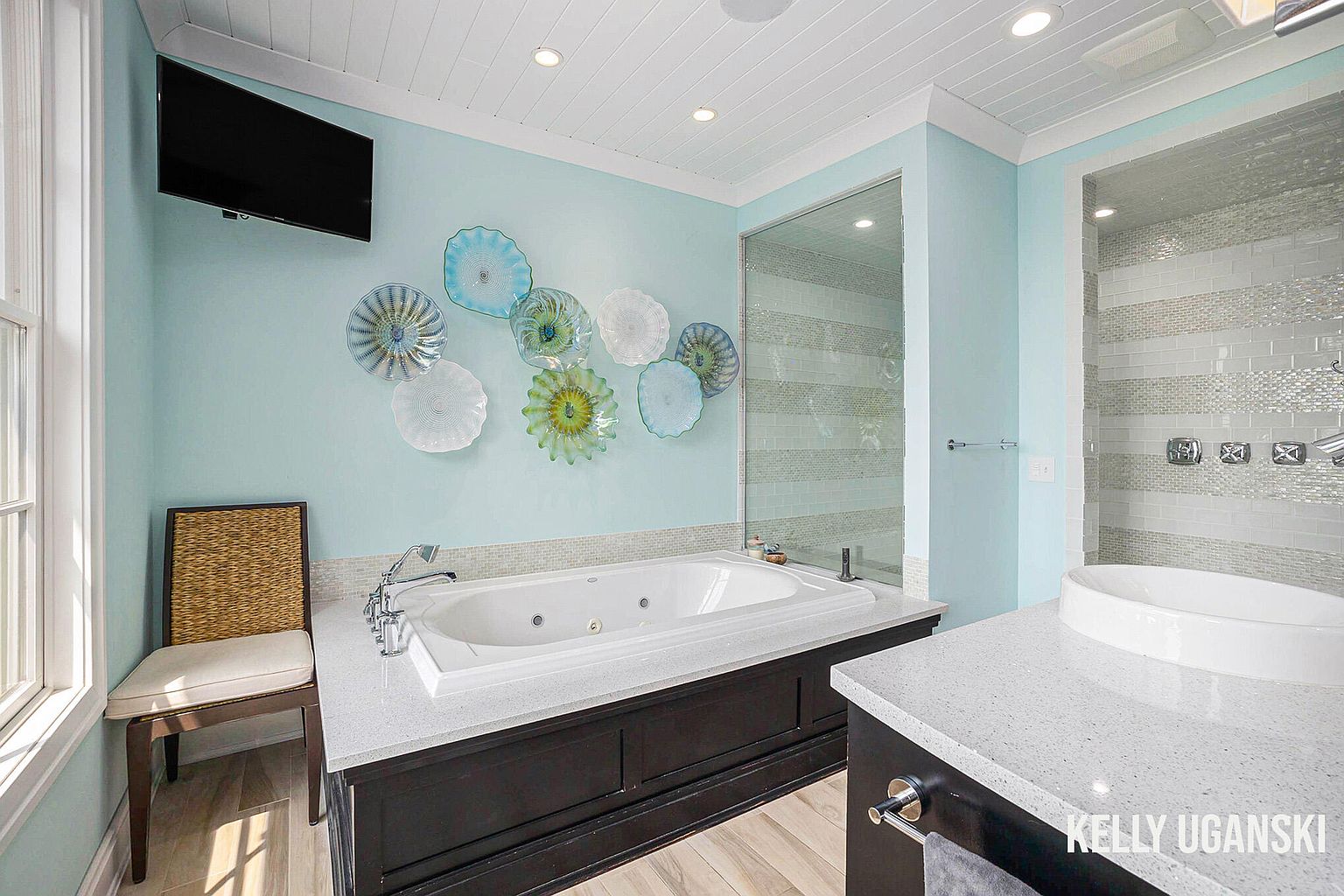 This is a primary bathroom featuring a large soaking tub with a white countertop and dark cabinetry. The walls are a calming light blue, adorned with decorative glass art. A walk-in shower with horizontally striped tile is visible, along with a separate vanity area. A chair sits near the window, and a flat-screen TV is mounted on the wall.
