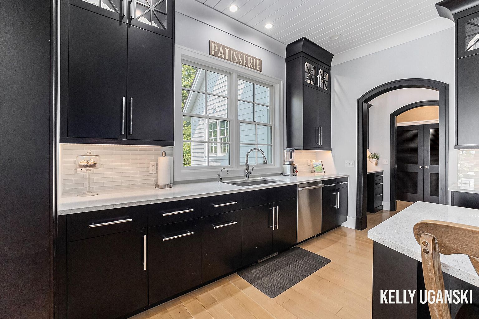 This is a well-lit kitchen featuring dark cabinetry with silver hardware, white countertops, and a white subway tile backsplash. A window above the sink provides natural light, and stainless steel appliances add a modern touch. The hardwood flooring and 'Patisserie' sign above the window add warmth and character to the space.